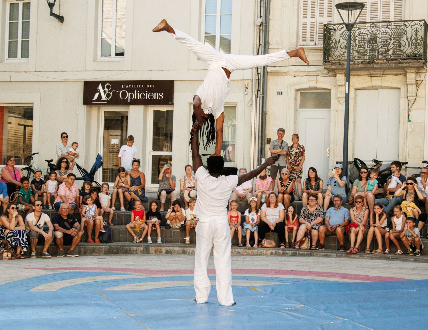 Acrobats perform a balancing act in front of a crowd on a blue performance area in a European street.