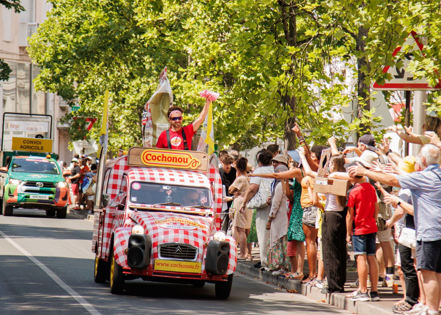 Red and white checkered vintage car drives through street parade as spectators cheer from sidewalk.