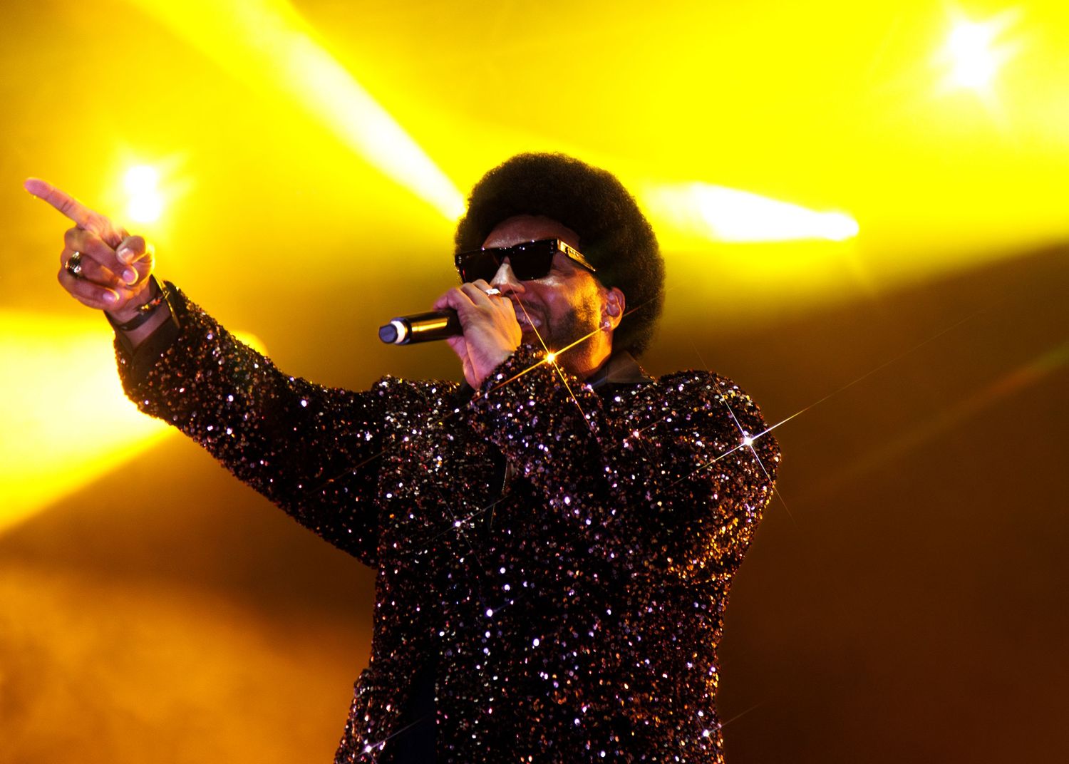 A performer in a sparkly black outfit performs on stage against a golden yellow backdrop.