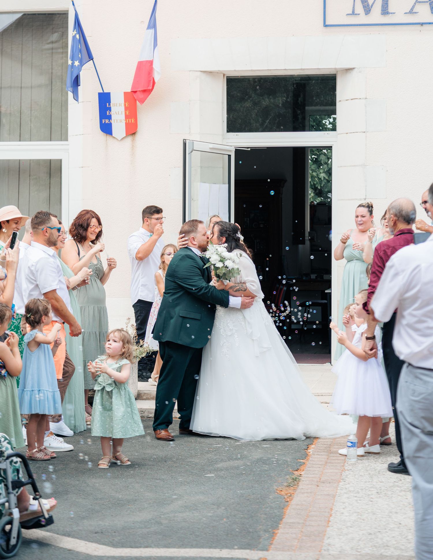 Newlyweds share first kiss outside French mairie surrounded by guests in sage green and white attire.