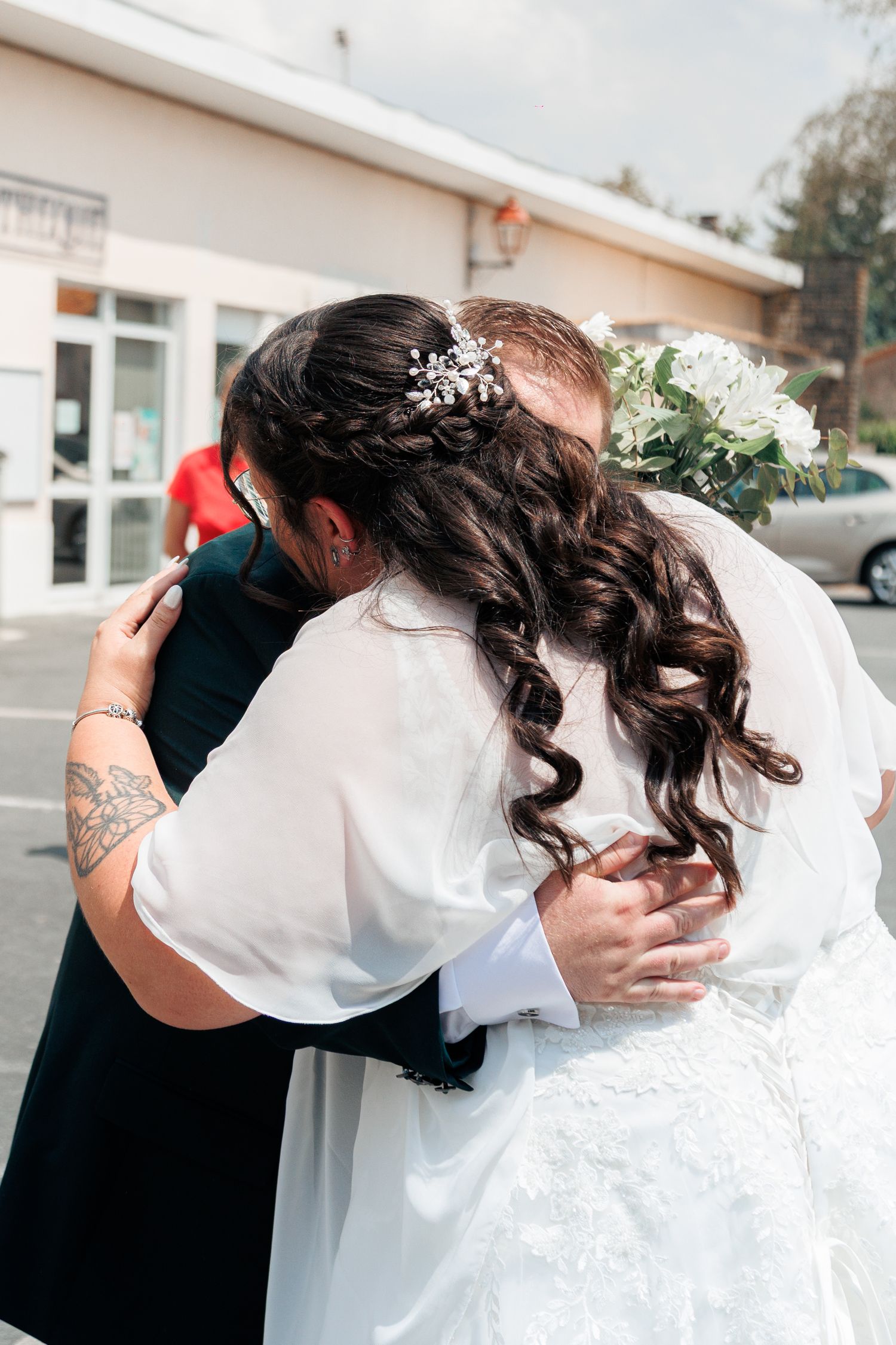 Bride and groom embracing outdoors on their wedding day, bride holding white bouquet with floral hair accessory.