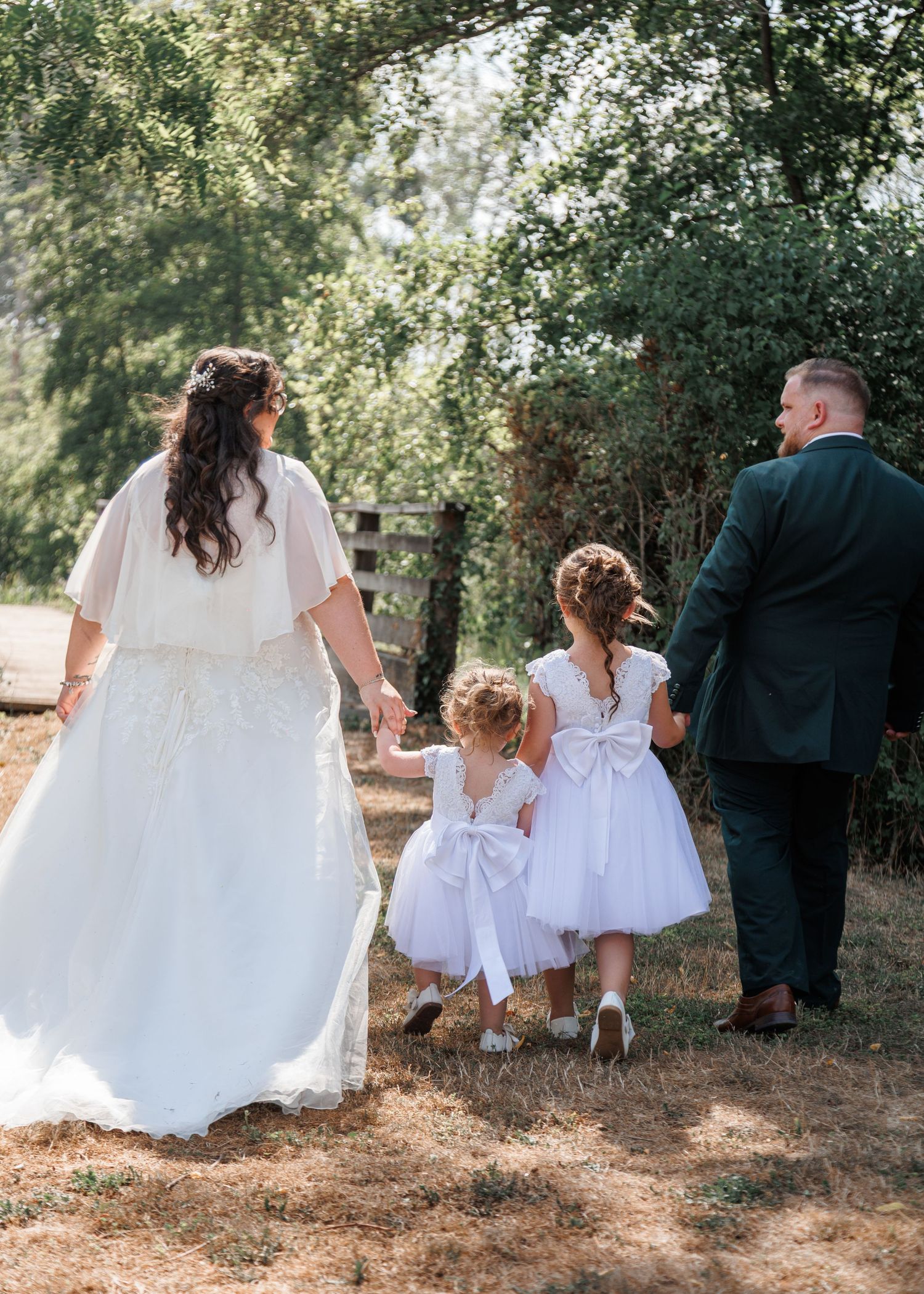 Bride and groom walk hand-in-hand with two flower girls in white dresses through a sunny garden.