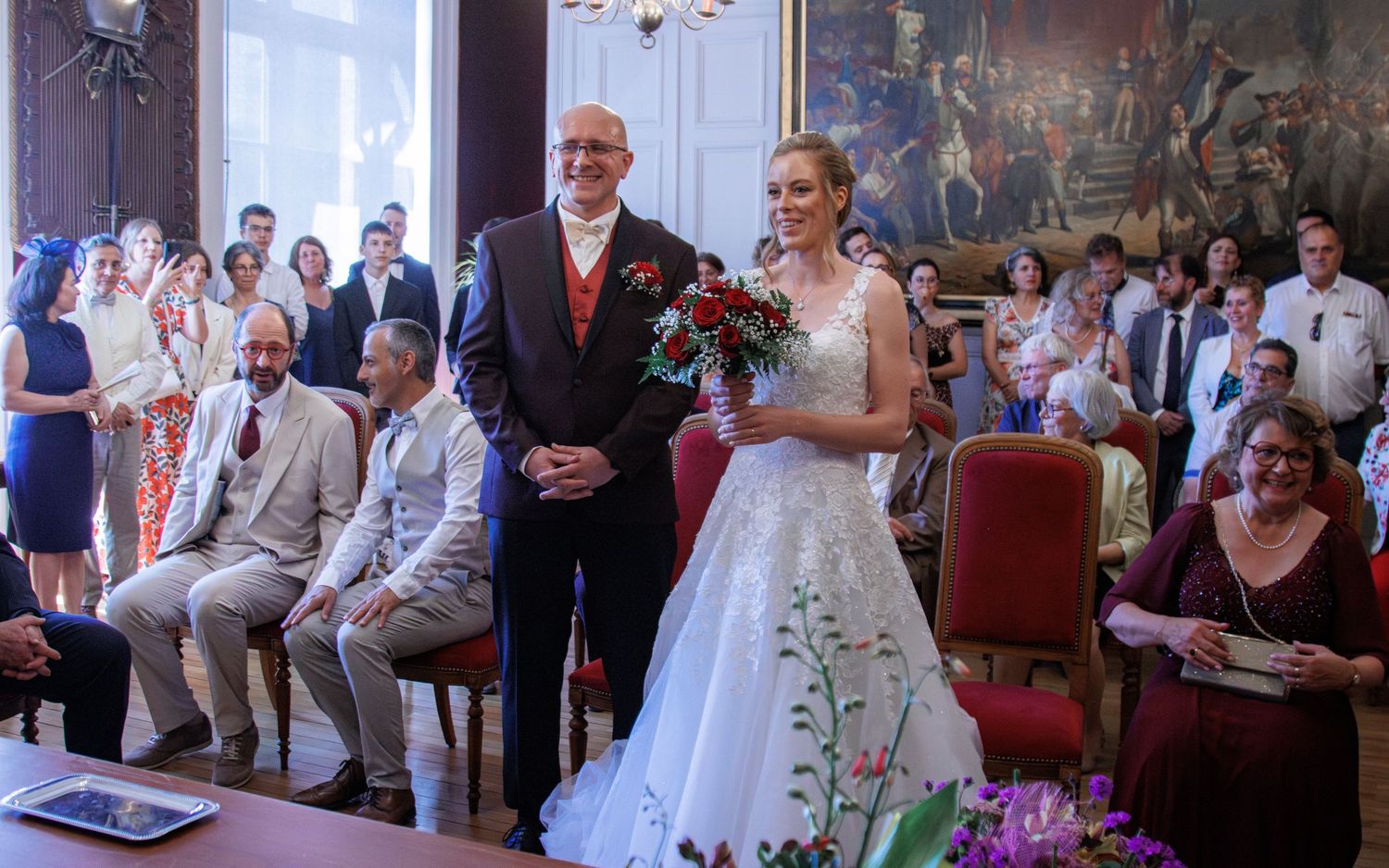 Bride in white floral gown holds red bouquet beside groom in wedding ceremony hall with seated guests.