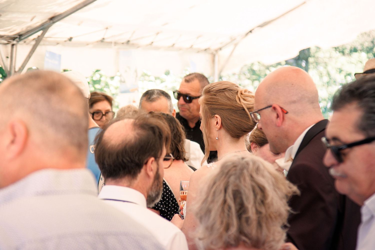 Wedding guests mingle under white tent canopy during outdoor wedding reception on a bright sunny day.
