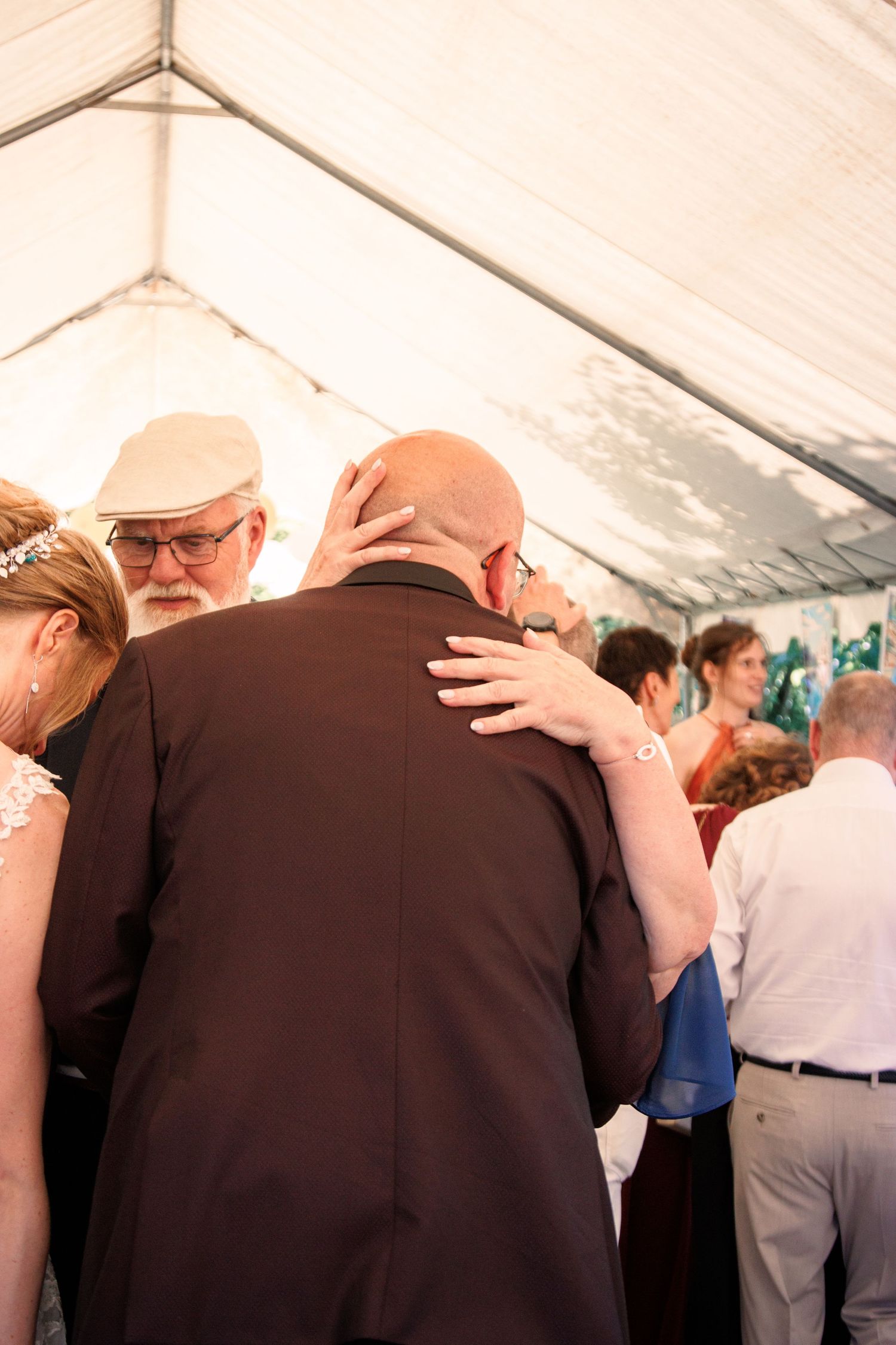 Guests mingling under a white tent at a wedding reception, with a bald man in a brown suit being embraced.