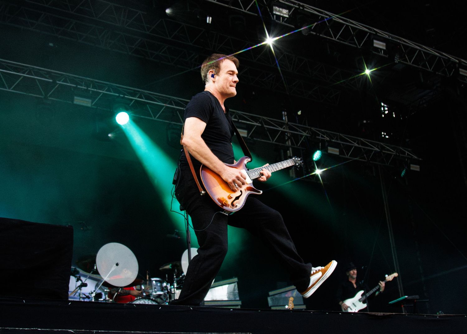 Guitarist performing energetically on stage under green lights at an outdoor concert festival.