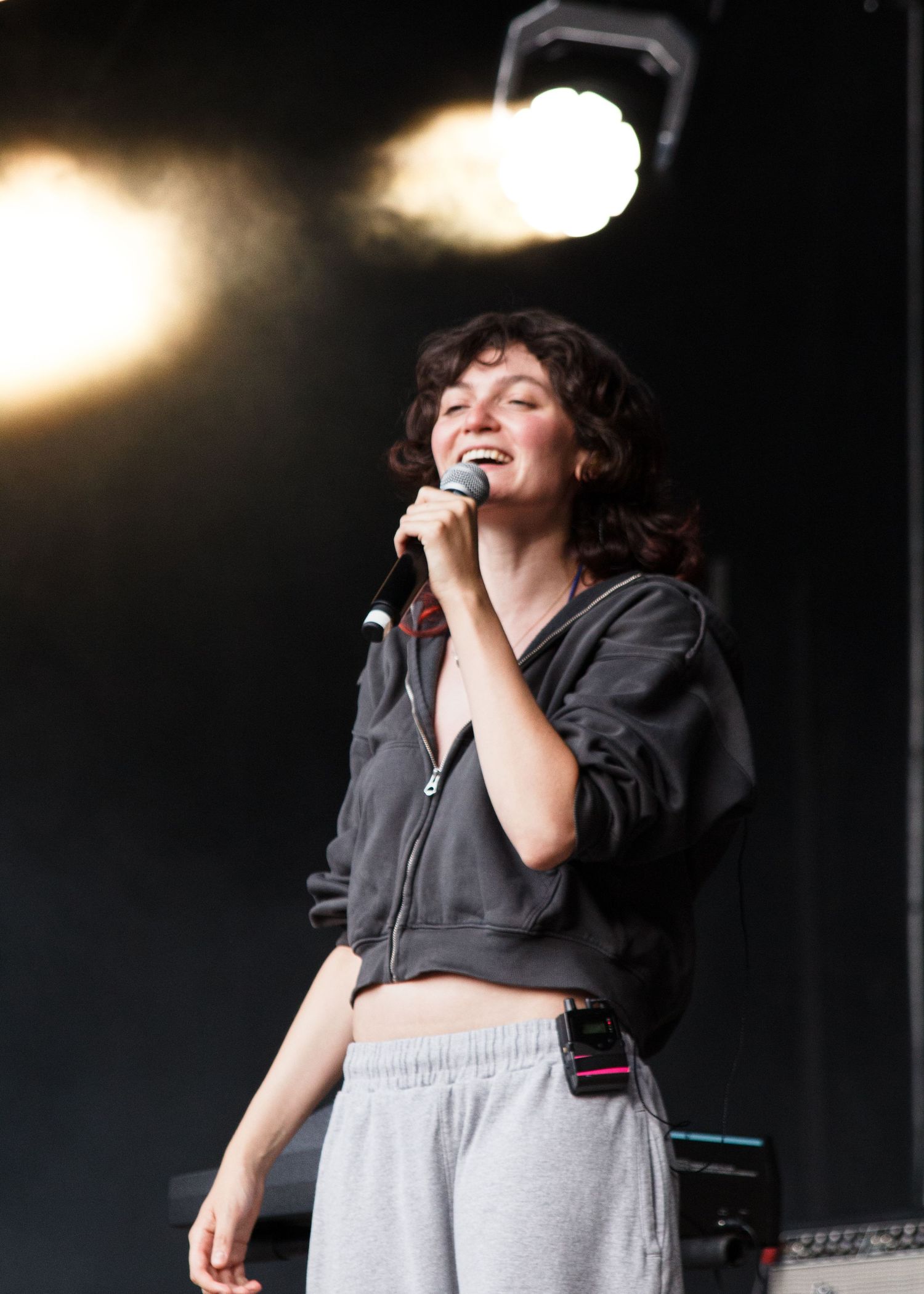 Young woman with curly hair singing into a microphone on stage, wearing a dark hoodie and grey sweatpants under bright stage lights.