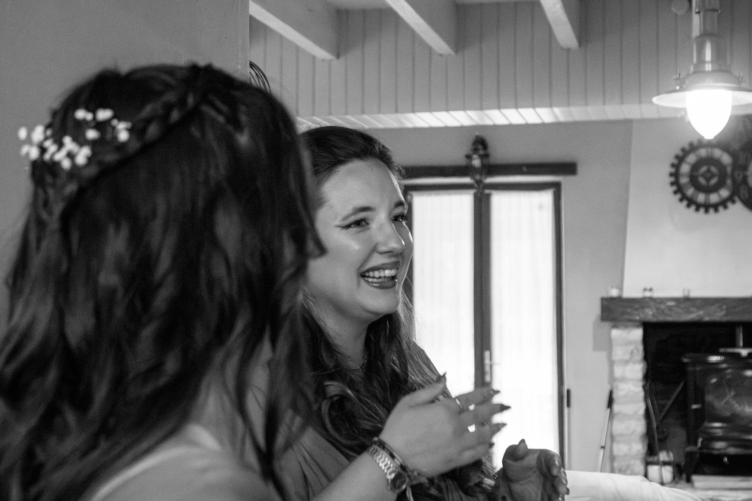 Two women share a joyful moment in a rustic interior, one wearing a floral crown, captured in black and white.