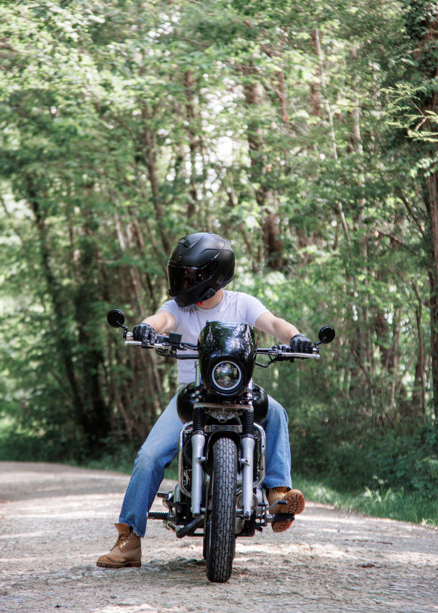 Motorcyclist wearing helmet and jeans rides a classic motorcycle on a tree-lined dirt road.