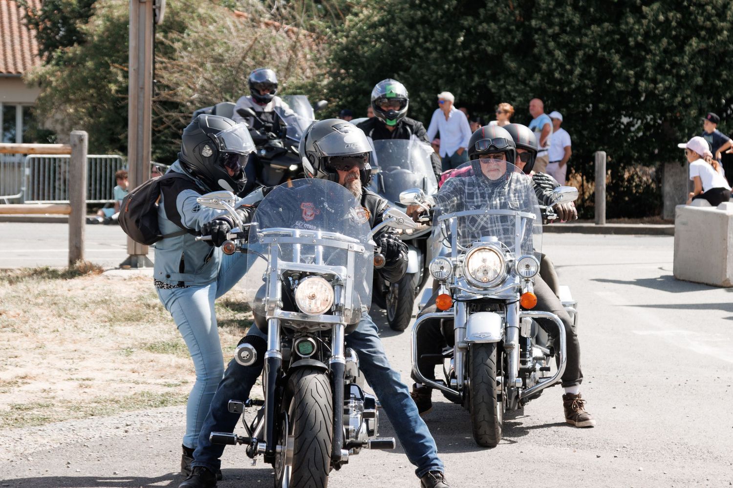 Group of motorcyclists riding Harley-Davidson bikes through a sunny street, wearing helmets and jeans.