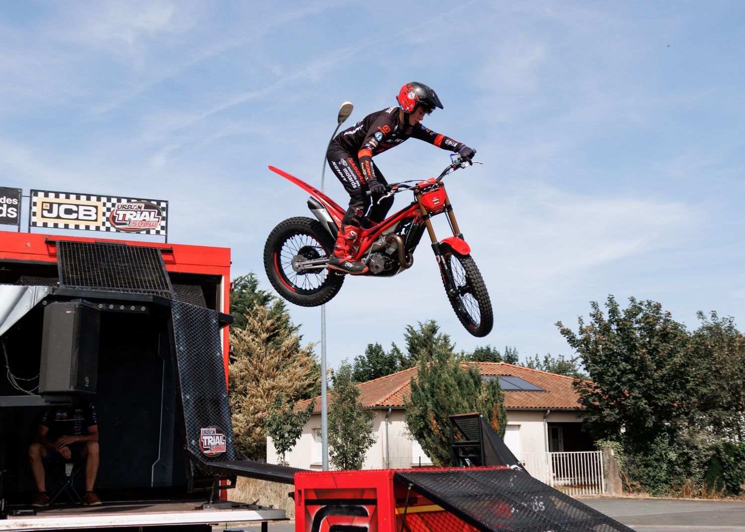 Motocross rider performing a ramp jump on a red dirt bike at an outdoor stunt event under a clear blue sky.