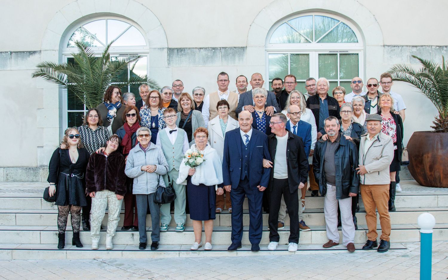 Large wedding group photo on building steps with bride and groom centered among formally dressed guests outdoors.