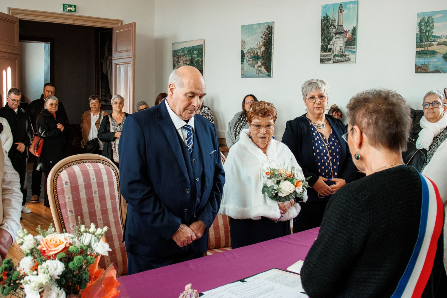 Elderly couple exchanging vows at a civil wedding ceremony with guests and an officiant at a decorated table.