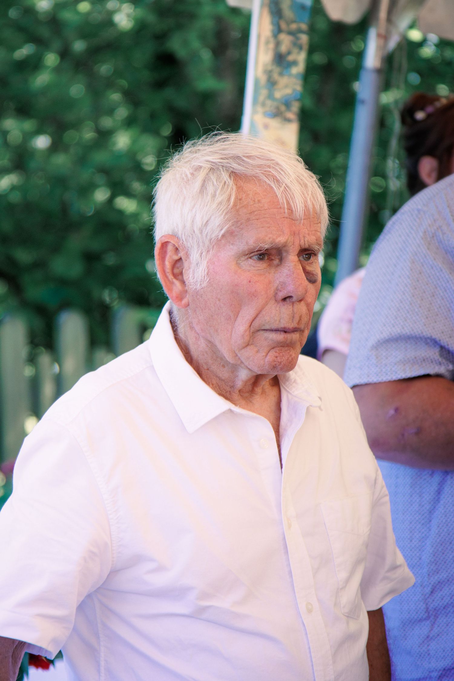 Elderly man with white hair wearing a white shirt standing outdoors with green trees in the background.