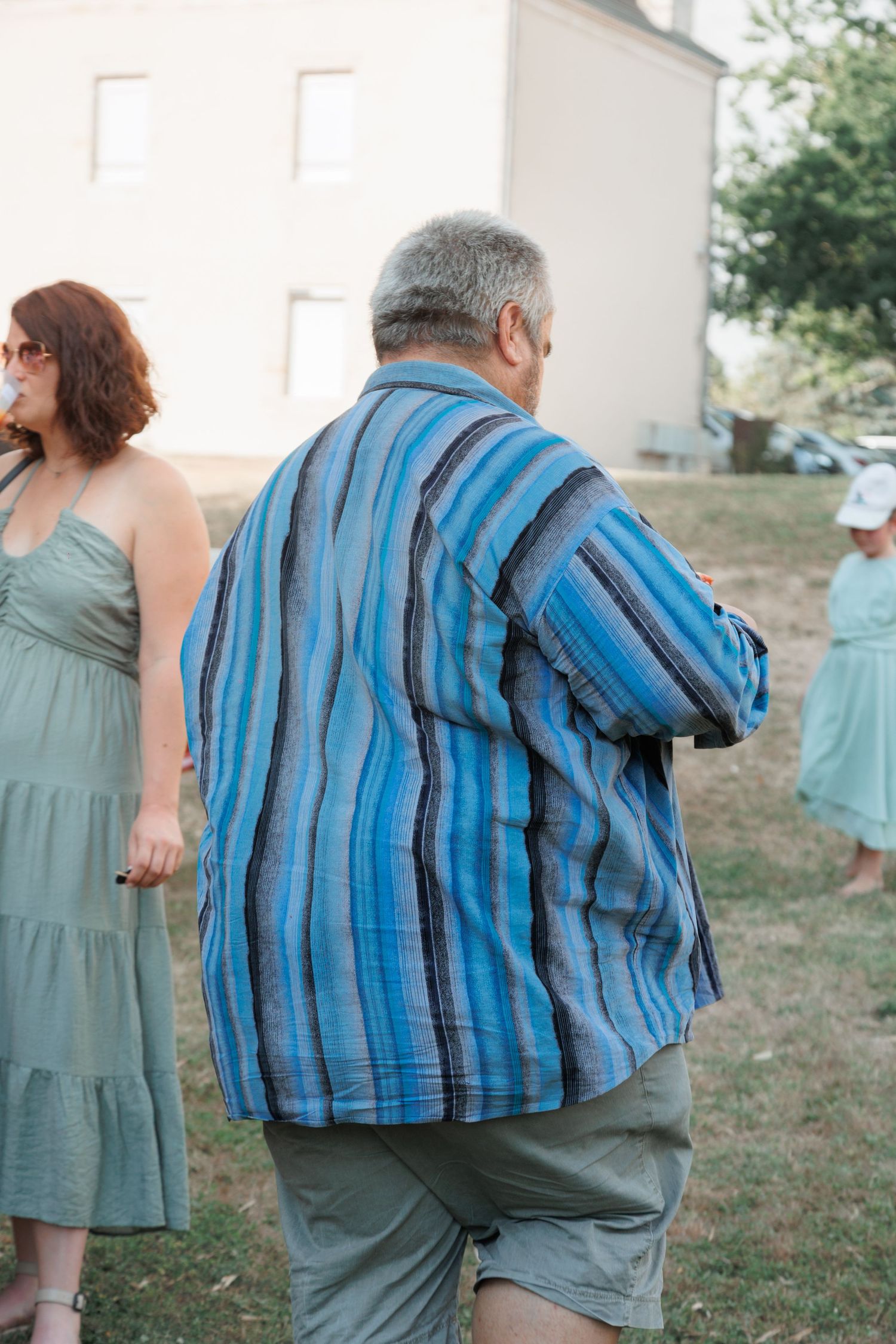 Older man in blue striped shirt stands outdoors at a wedding celebration with guests nearby.