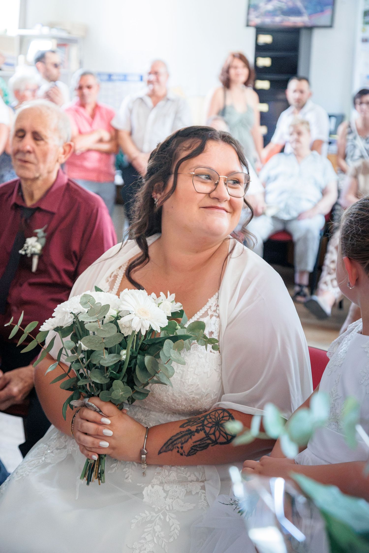 Bride in white lace gown holding white flower bouquet smiles during indoor wedding ceremony.