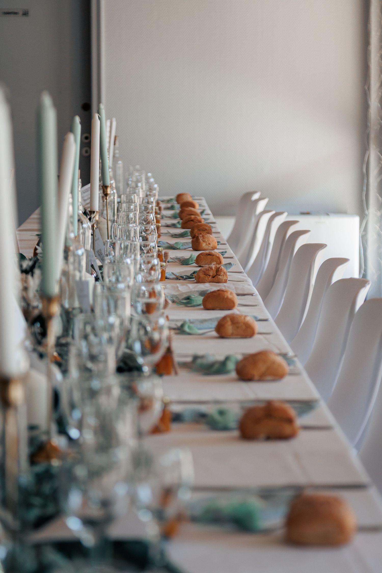 Long elegantly set wedding reception table with white chairs, candles, glassware, and bread rolls.