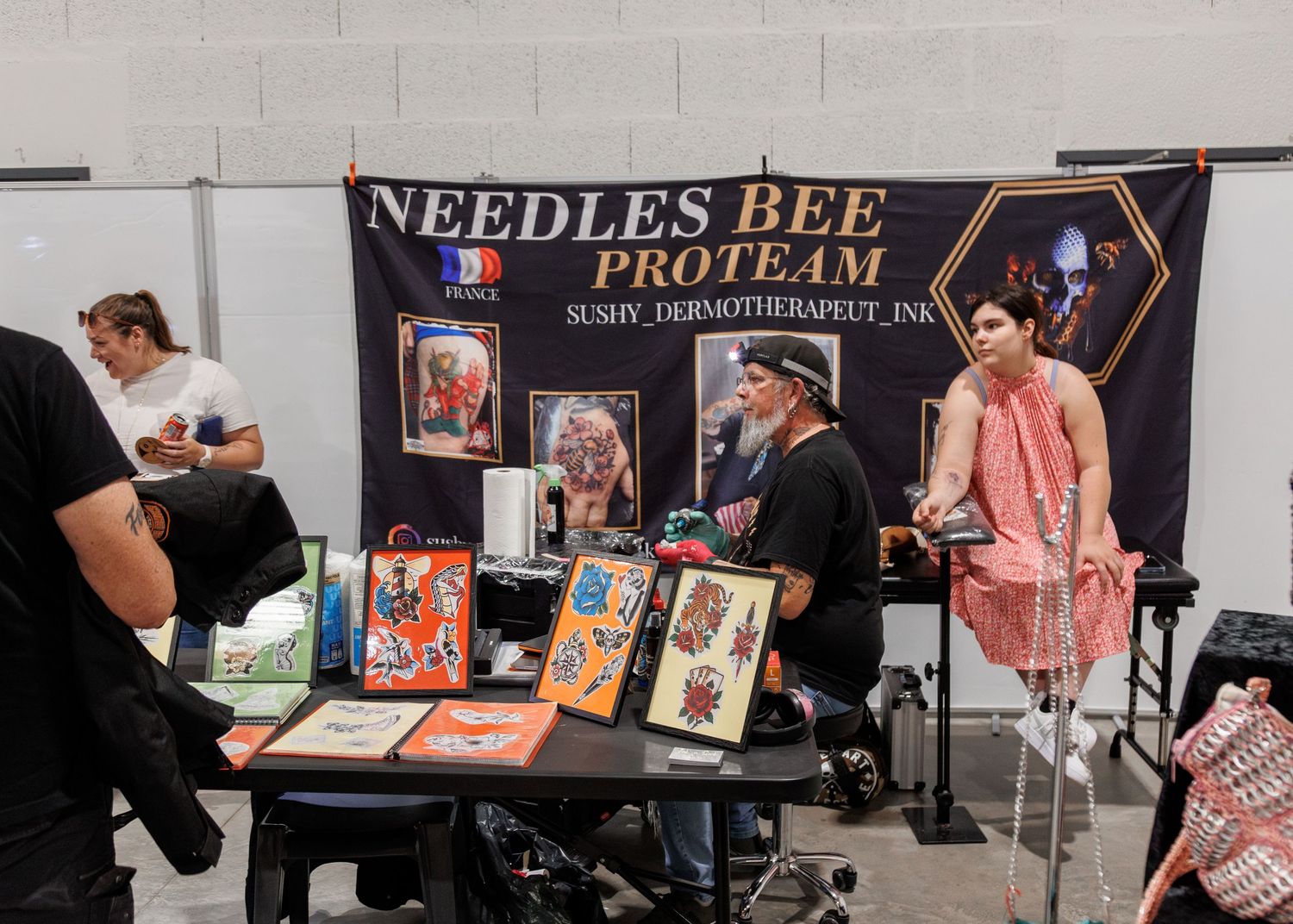 Needles Bee Proteam tattoo convention booth displaying artwork and merchandise at an indoor tattoo expo.