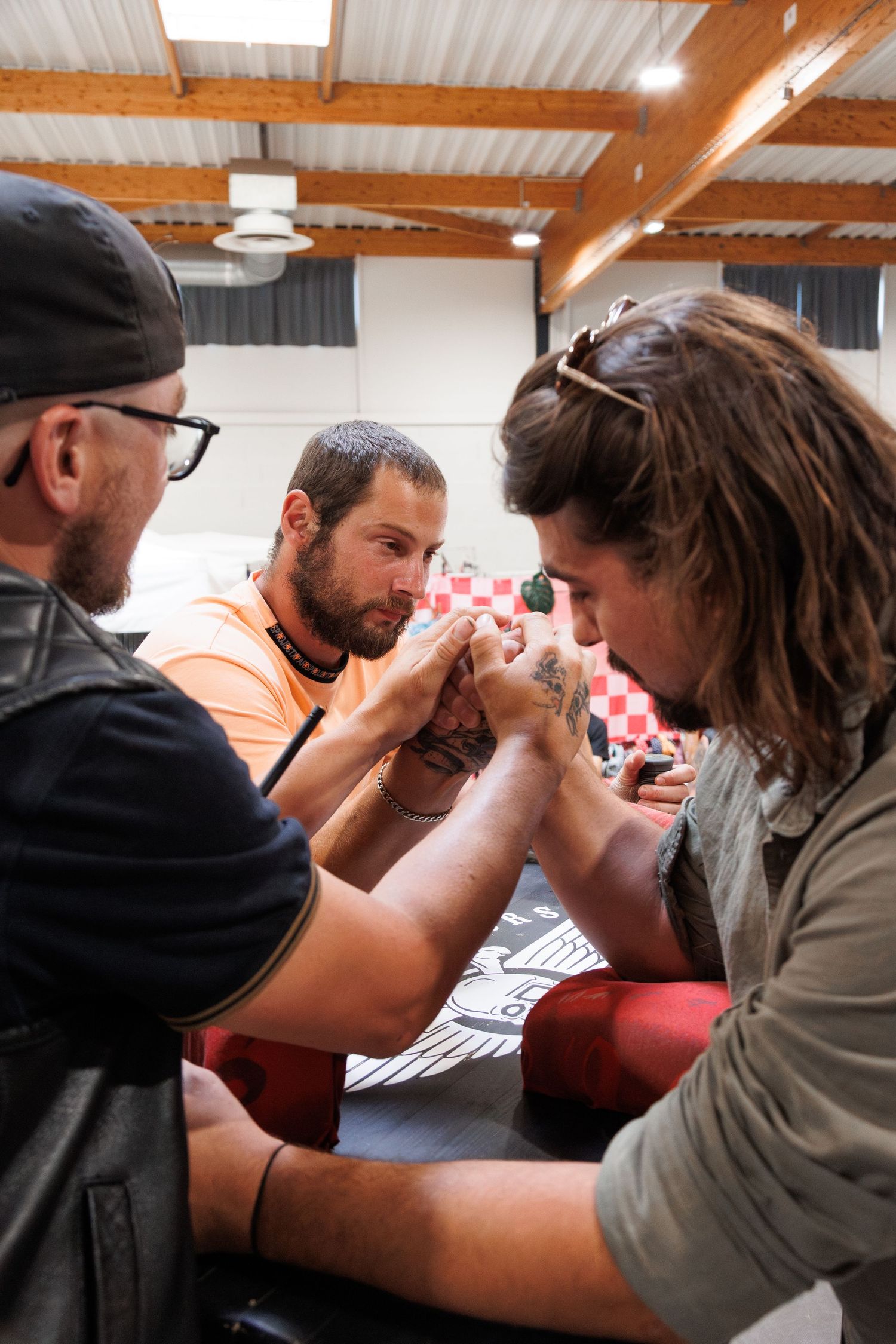 Two men arm wrestling at a table while a third man watches closely indoors at what appears to be a competition event.