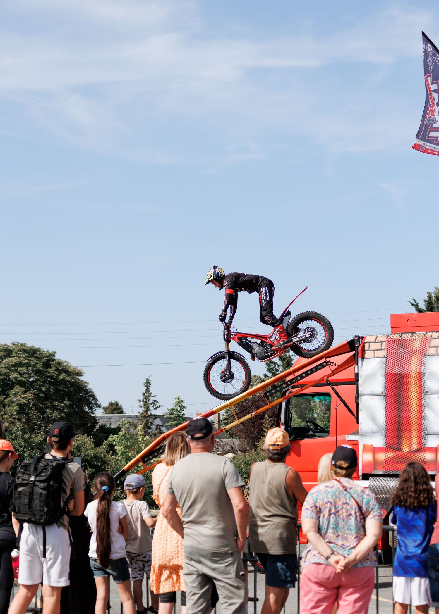 A motocross rider launches off a ramp at an outdoor stunt show while a crowd watches below near a red truck.