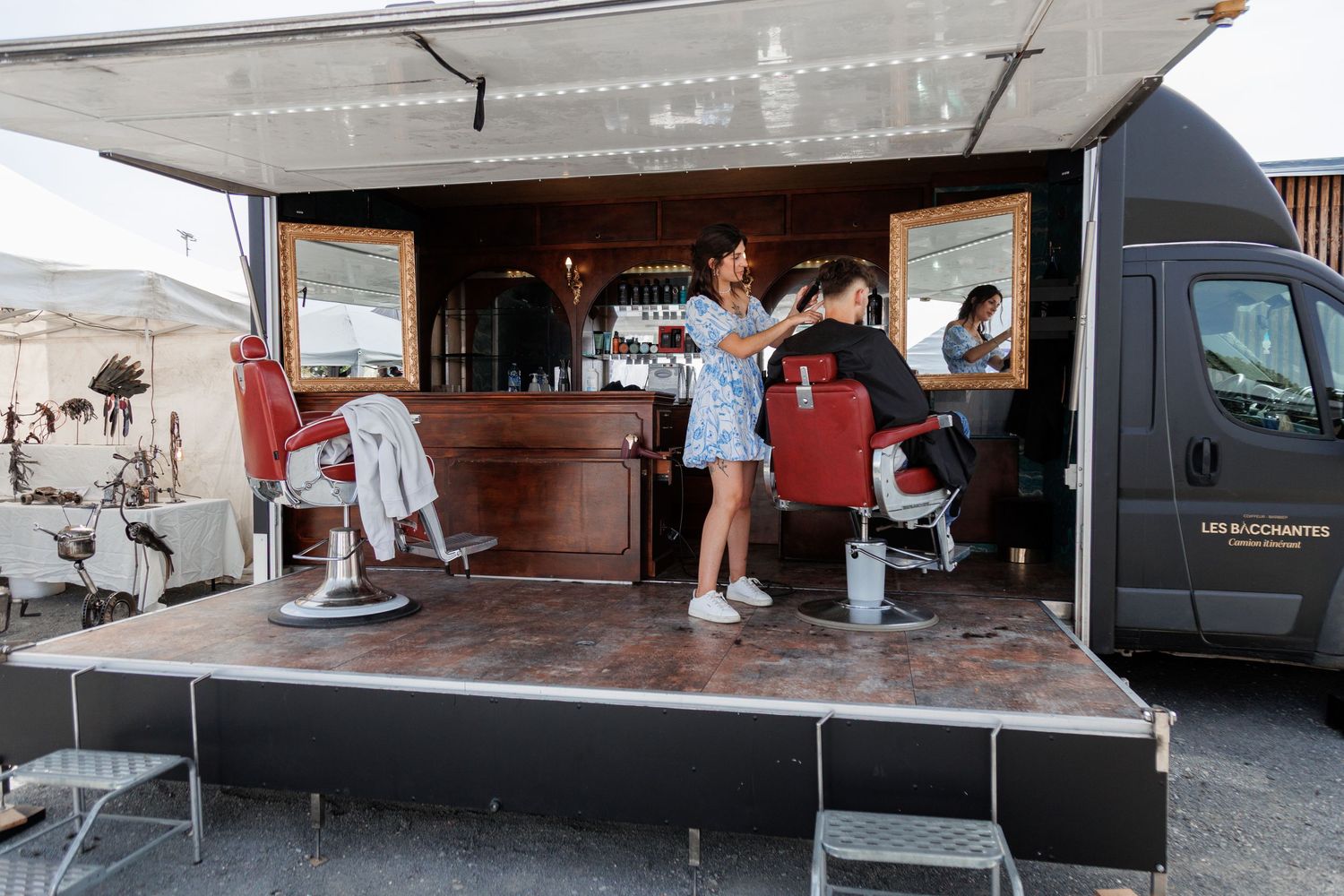 Mobile barber shop truck with two red chairs and wooden bar serving customers at outdoor event.