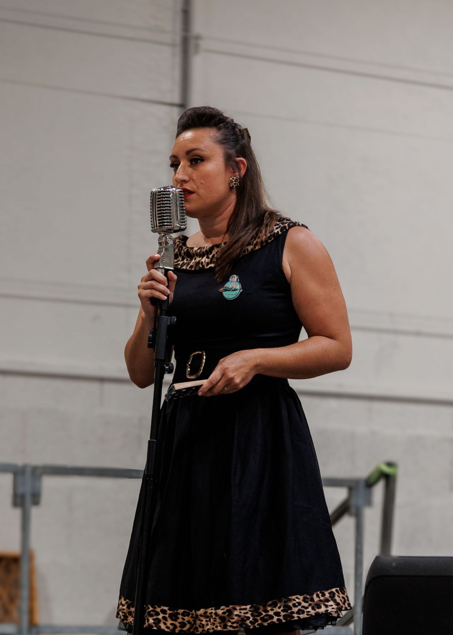 Woman in black leopard-trim dress singing into vintage microphone on stage.