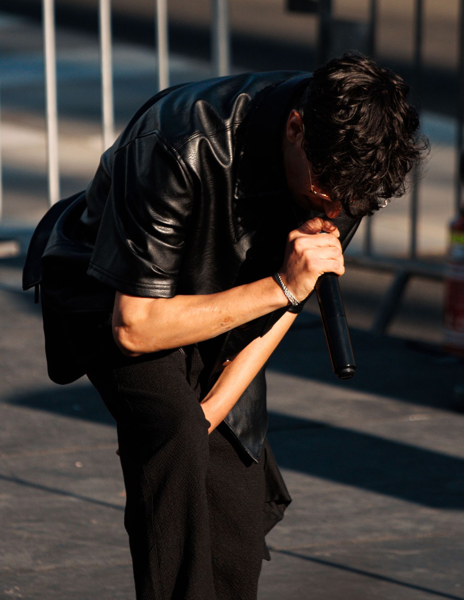 Performer in black leather jacket leaning forward with microphone on stage.