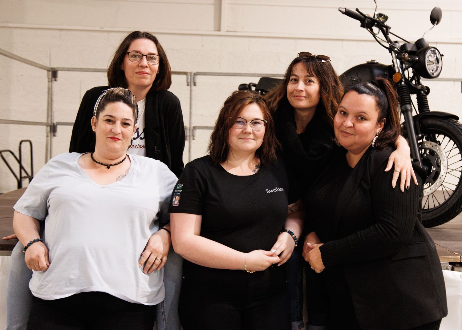 Friends gather for a casual group photo in front of a motorcycle in an indoor setting.