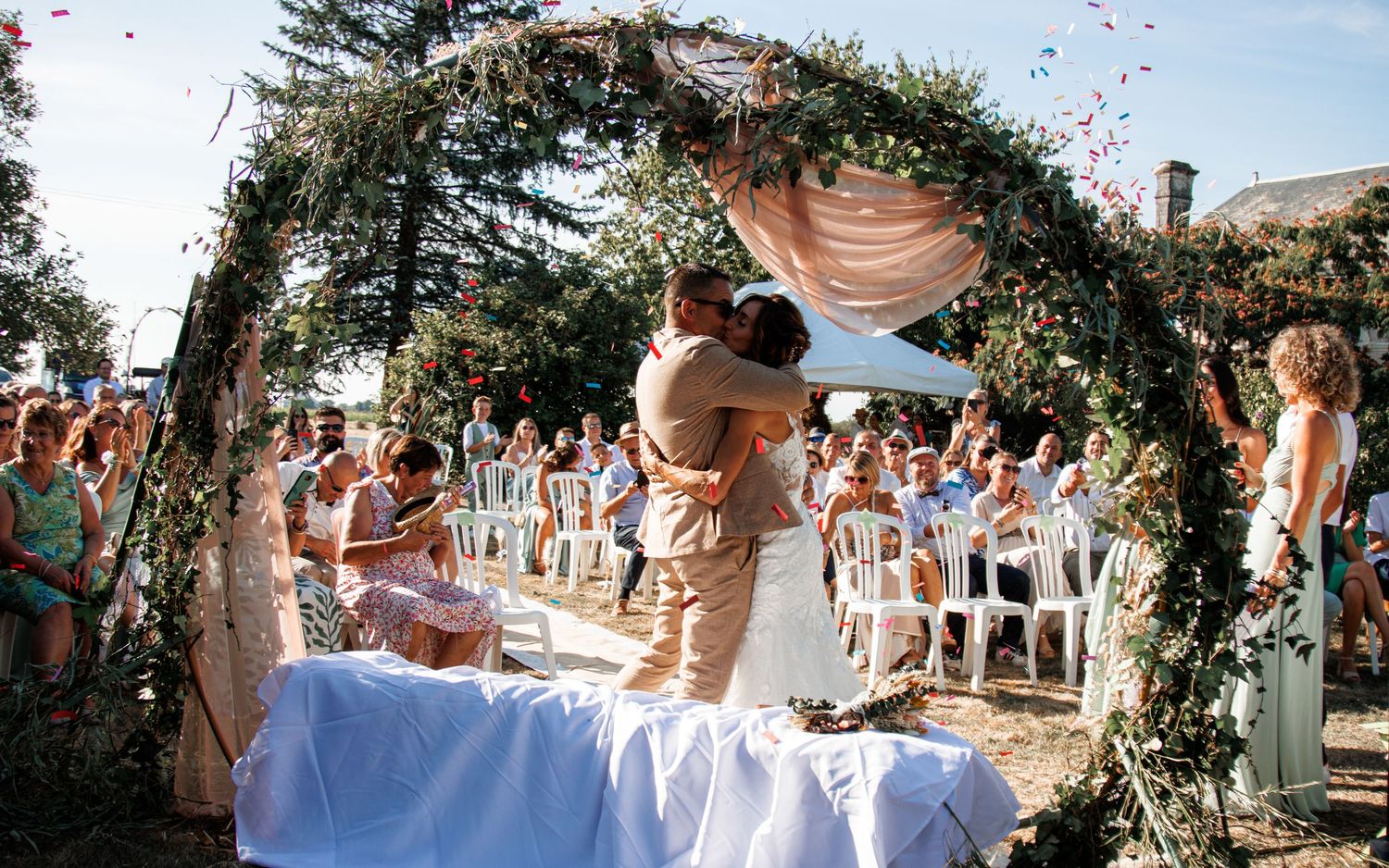 An intimate garden wedding ceremony takes place under a lush greenery arch with white draping and chairs on a sunny day.