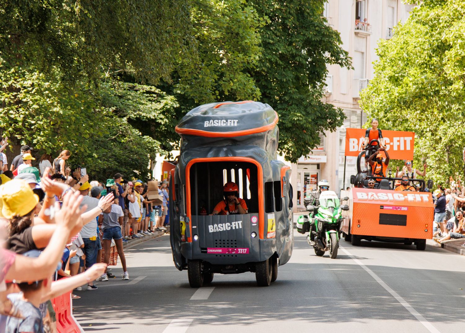 Decorated parade vehicles moving through tree-lined city street with cheering spectators.