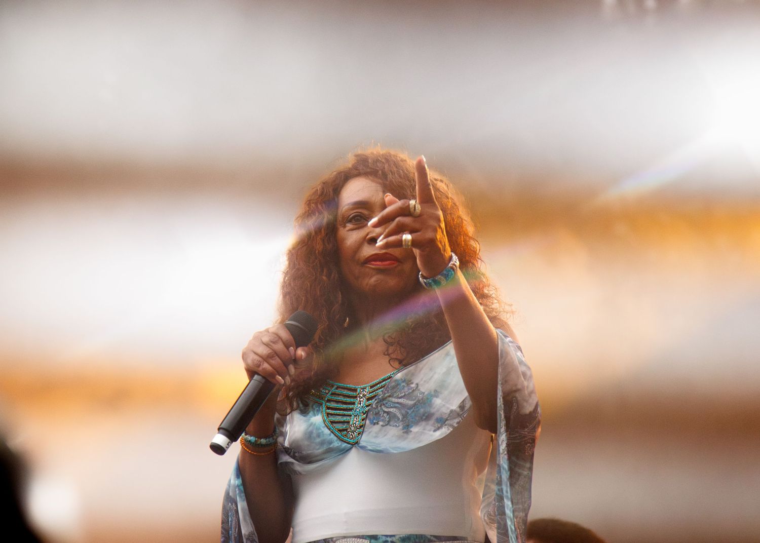 A performer singing on stage in a white top with warm sunset lighting and lens flare effects.