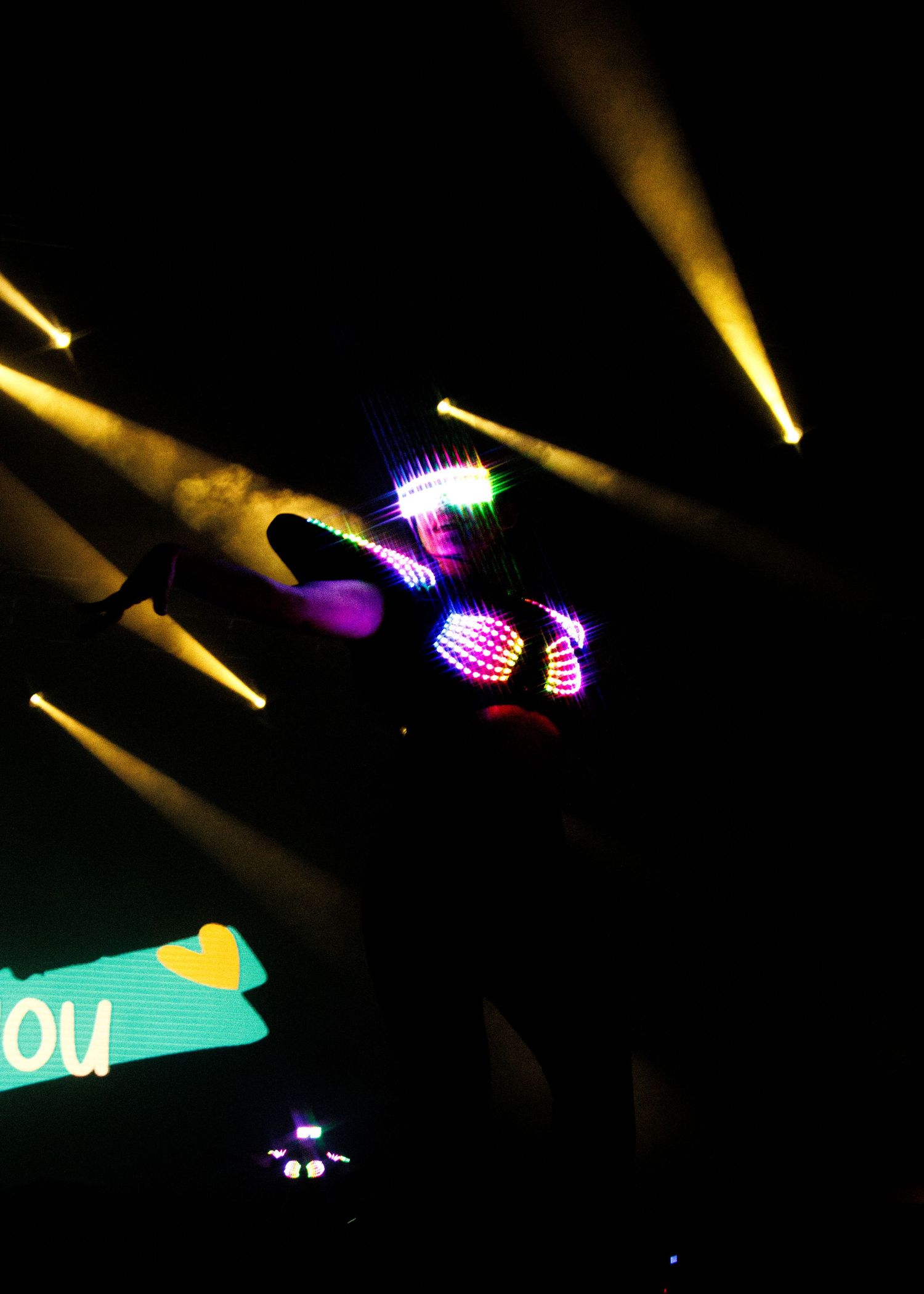 Colorful lights illuminate a dark concert or performance venue with neon purple and yellow beams shining in different directions.