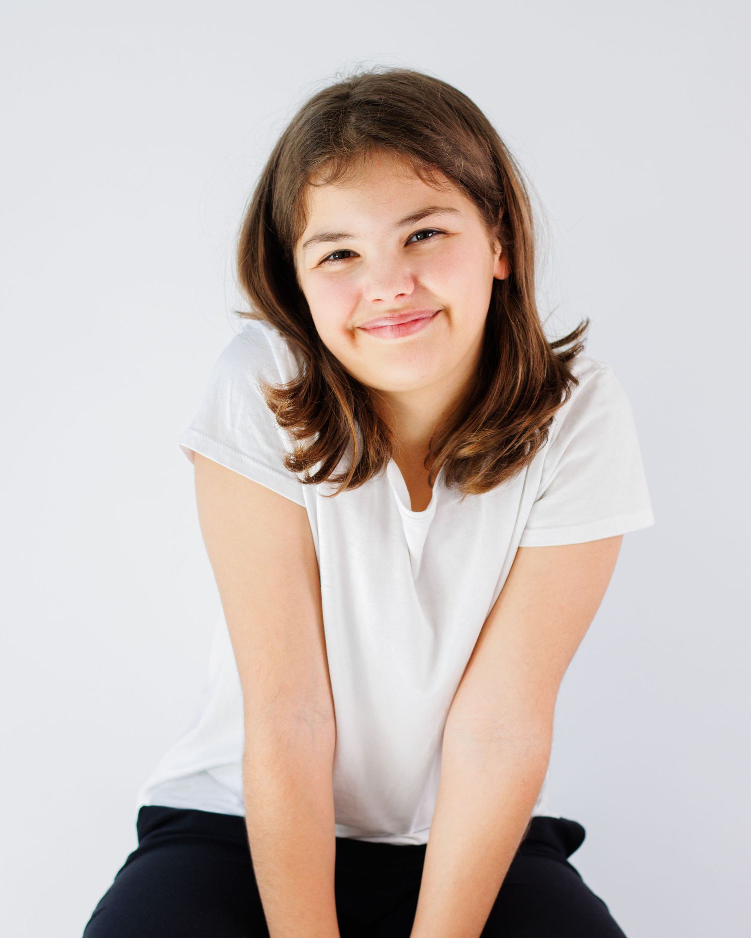 Person in white t-shirt and dark pants sitting casually while smiling warmly against light background.