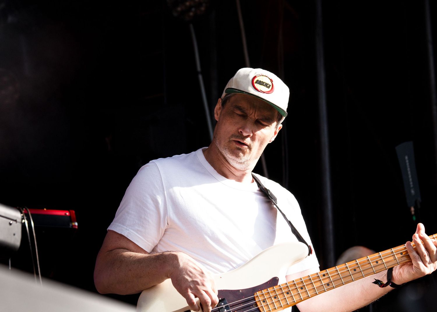 Bass player wearing a white t-shirt and baseball cap performs on stage during a live music performance.