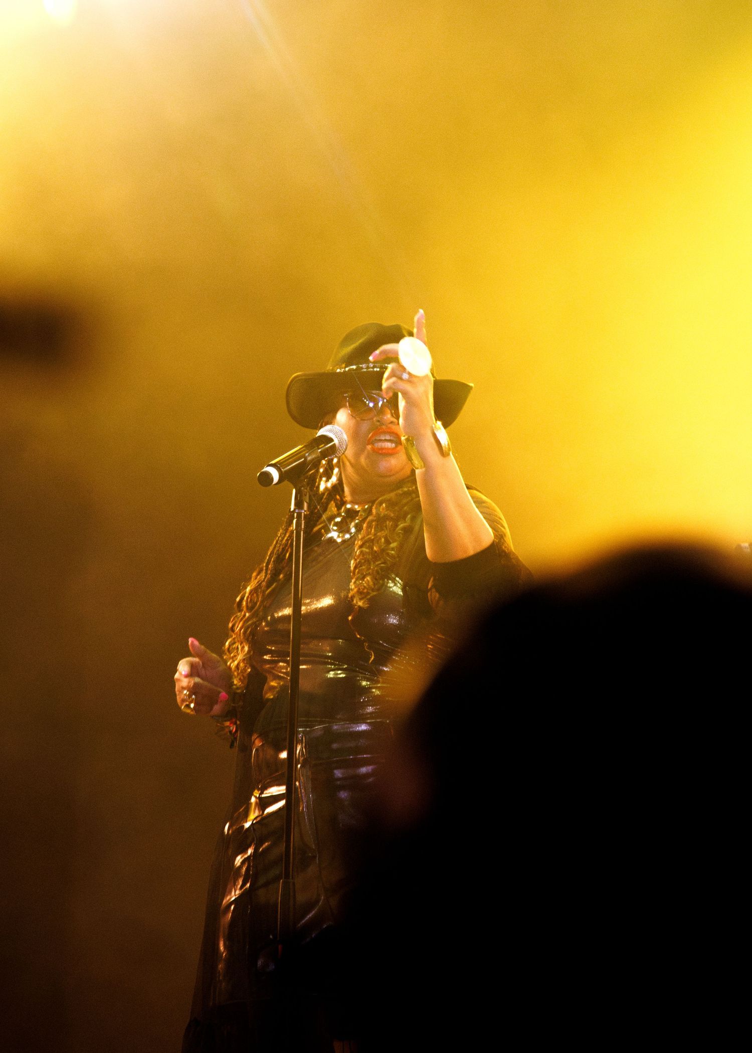 A silhouetted performer stands on stage with microphone against a bright golden yellow lighting backdrop.