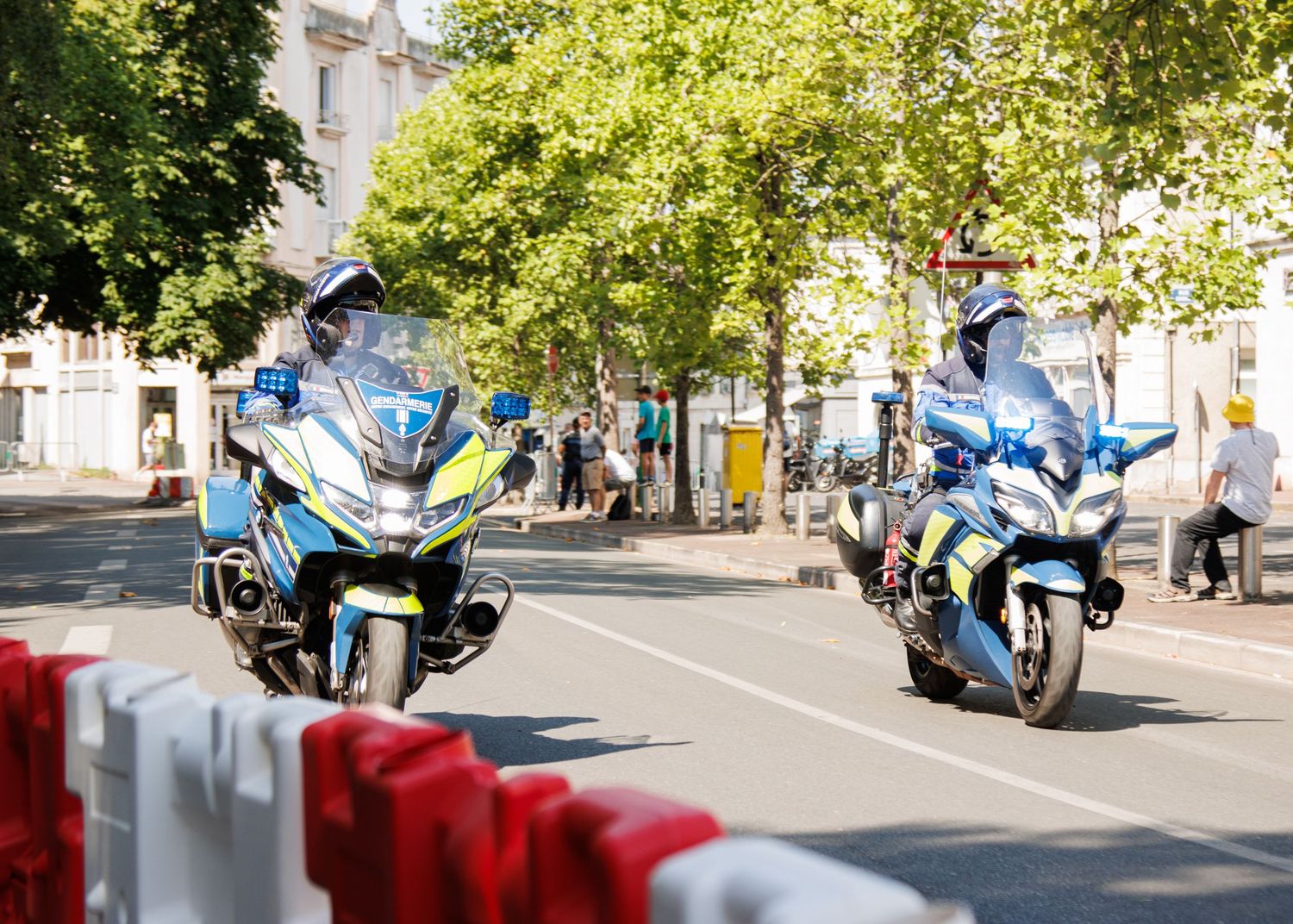 Performance motorcycles line up along a sunny tree-lined street during a parade or event.