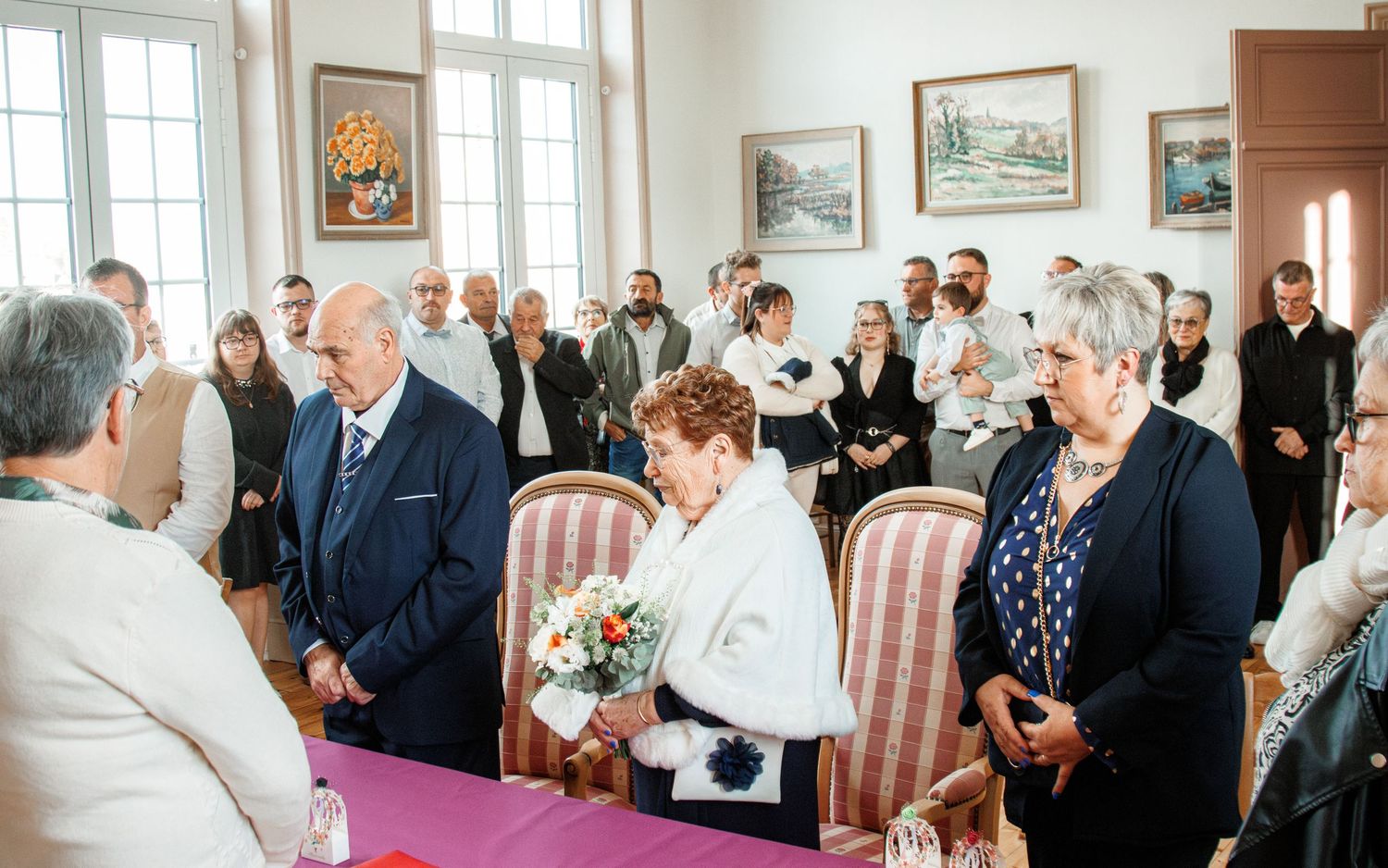 Elderly couple exchanging vows at an intimate civil wedding ceremony surrounded by guests in a decorated room.