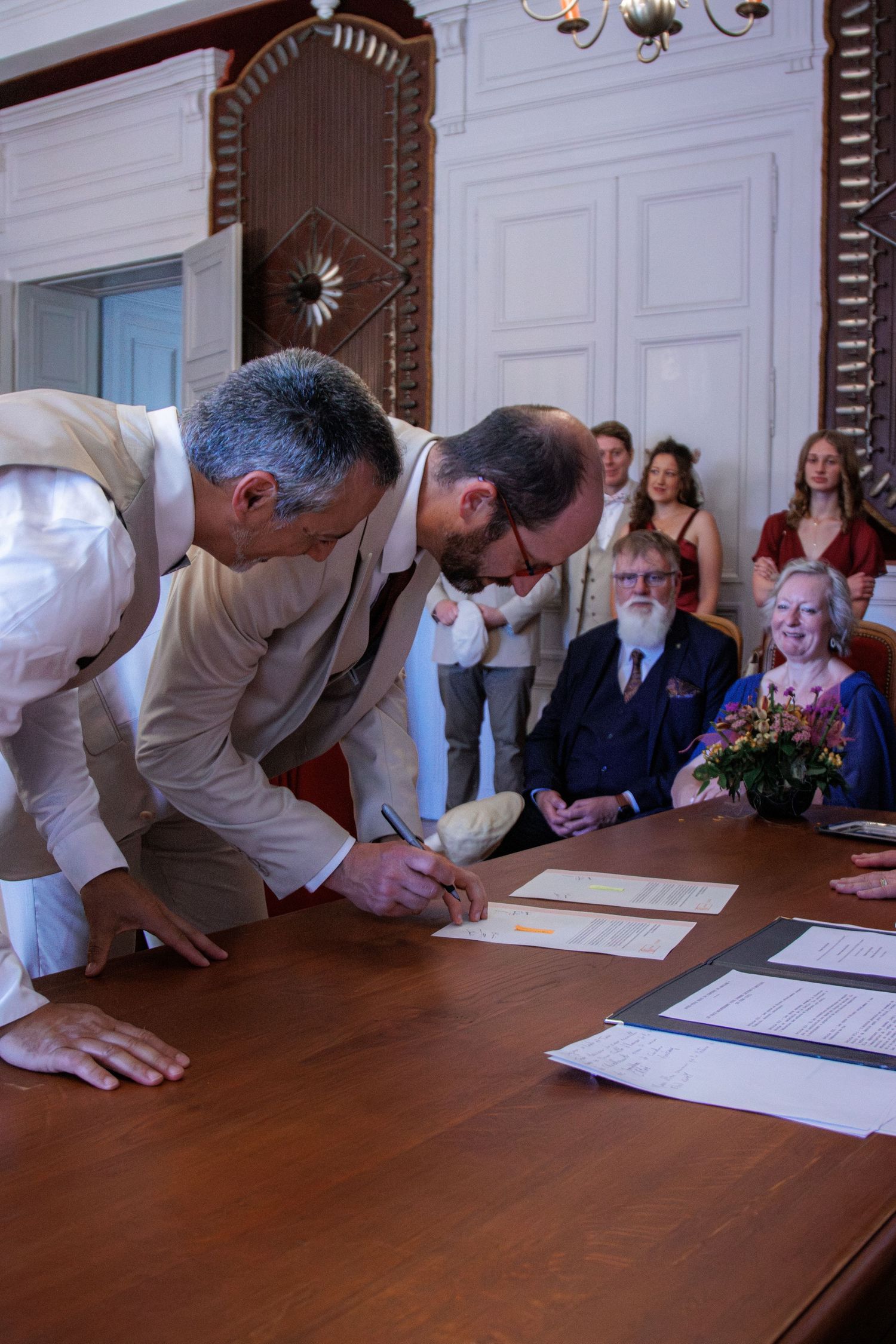 Two men signing wedding documents at a wooden table during a civil ceremony with guests watching in background.