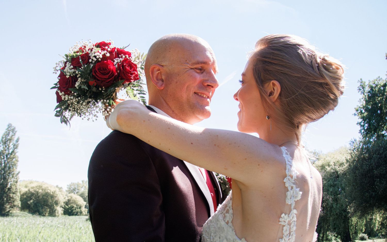 Smiling bald groom in burgundy suit gazes lovingly at bride holding red roses bouquet outdoors.