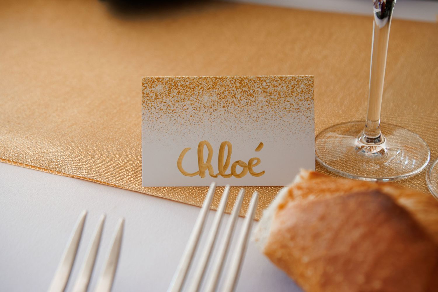 Elegant gold wedding place card with name Chloe beside champagne flute and fork.