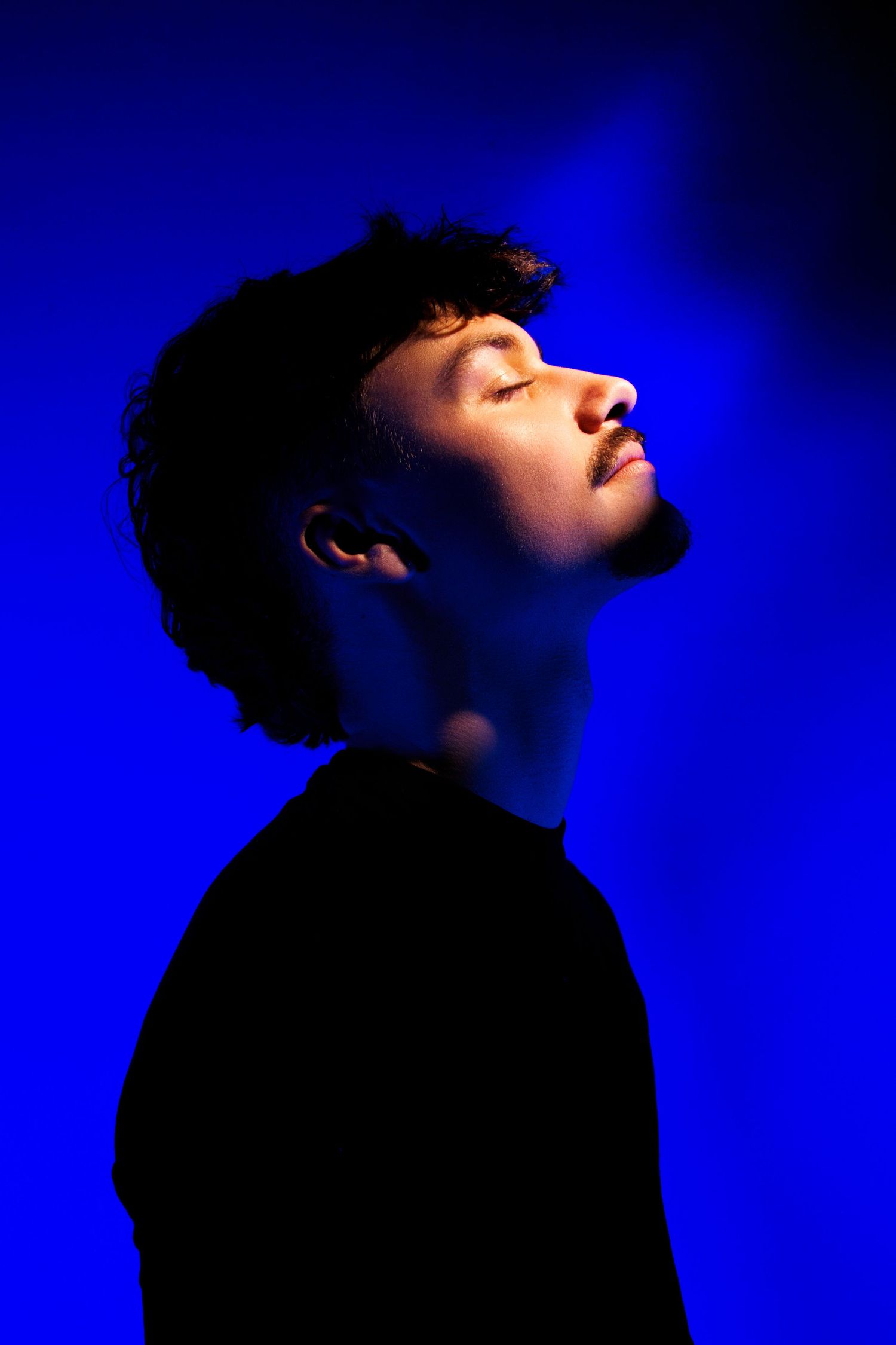 Young man with curly hair and closed eyes posed against a vivid blue background in dramatic studio lighting.