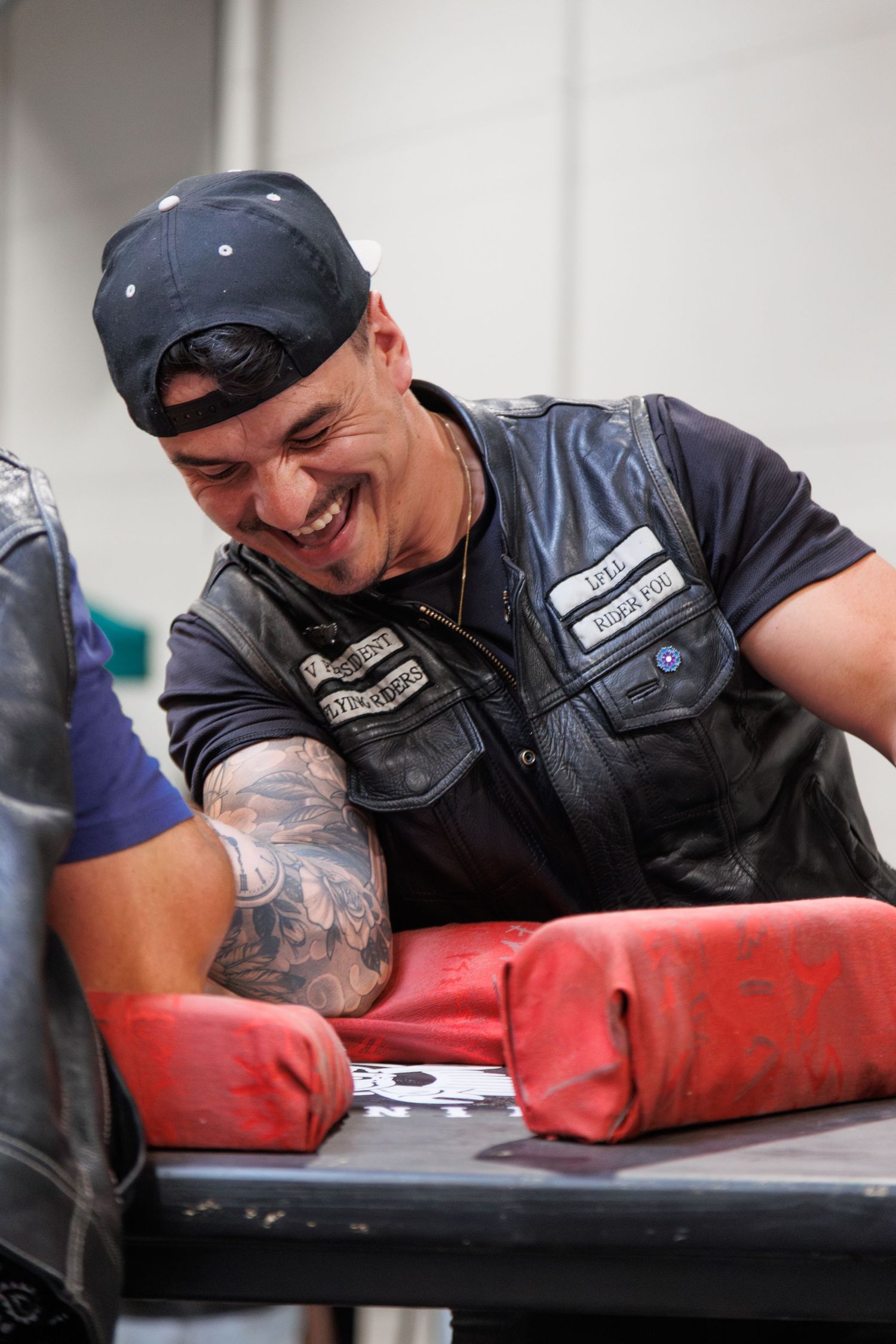 Smiling man in leather vest and backward cap with tattoos competing in arm wrestling at a table.