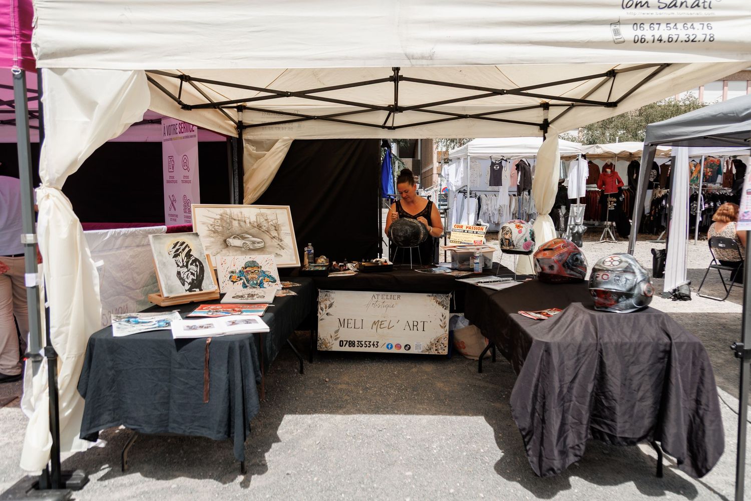 Artist vendor booth at outdoor market with artwork displays, dark tablecloths, and white canopy tent on sunny day.