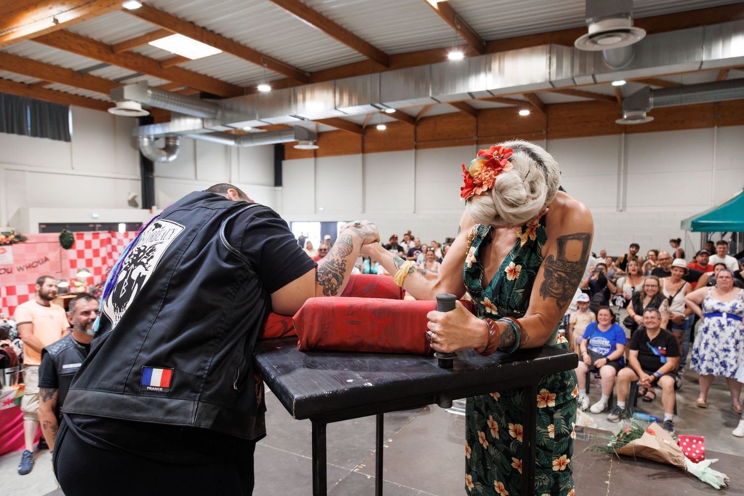 Two competitors engage in an arm wrestling match at an indoor tournament event with a crowd of spectators watching.