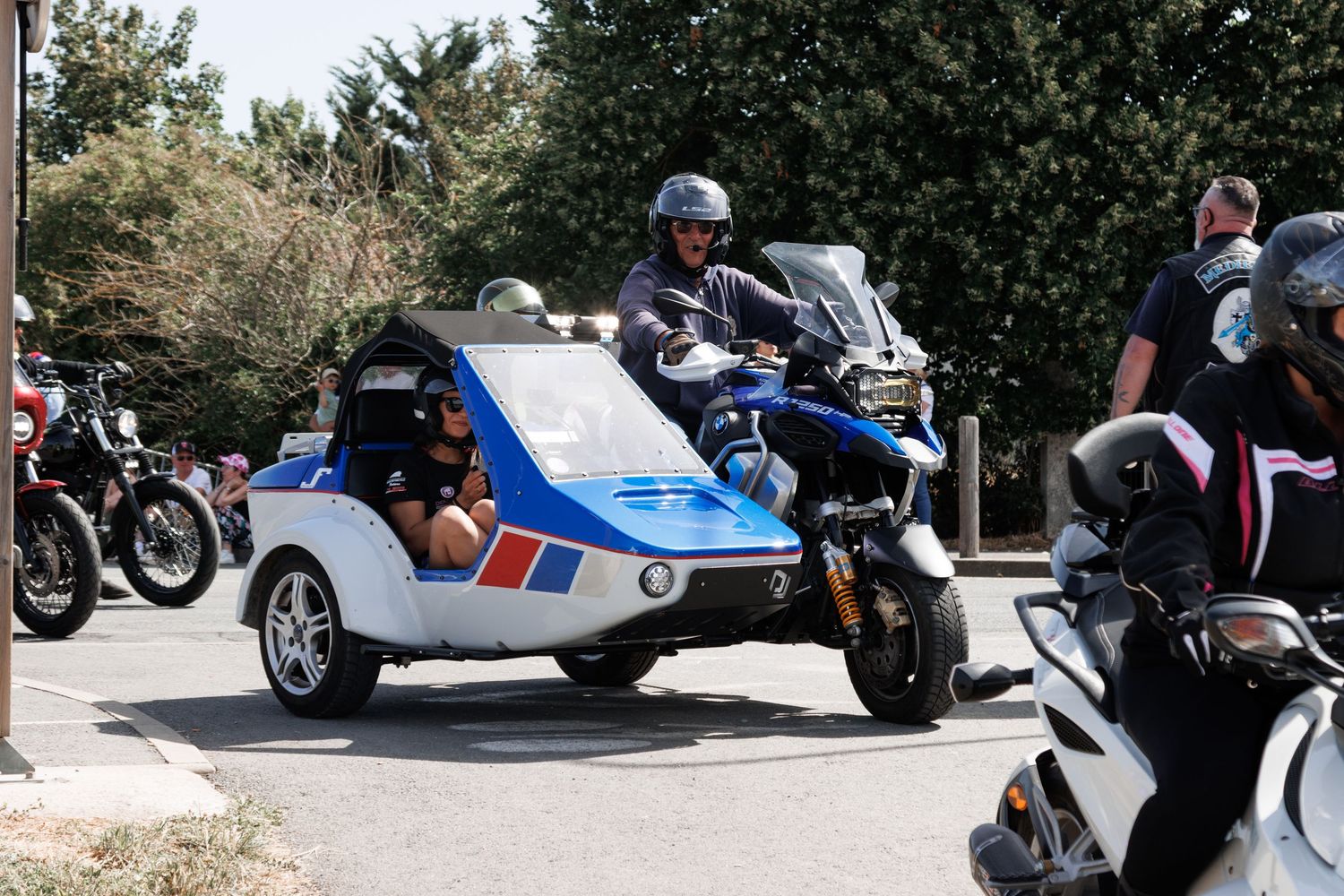 Police officer riding a blue and white motorcycle with sidecar at an outdoor motorcycle rally on a sunny day.