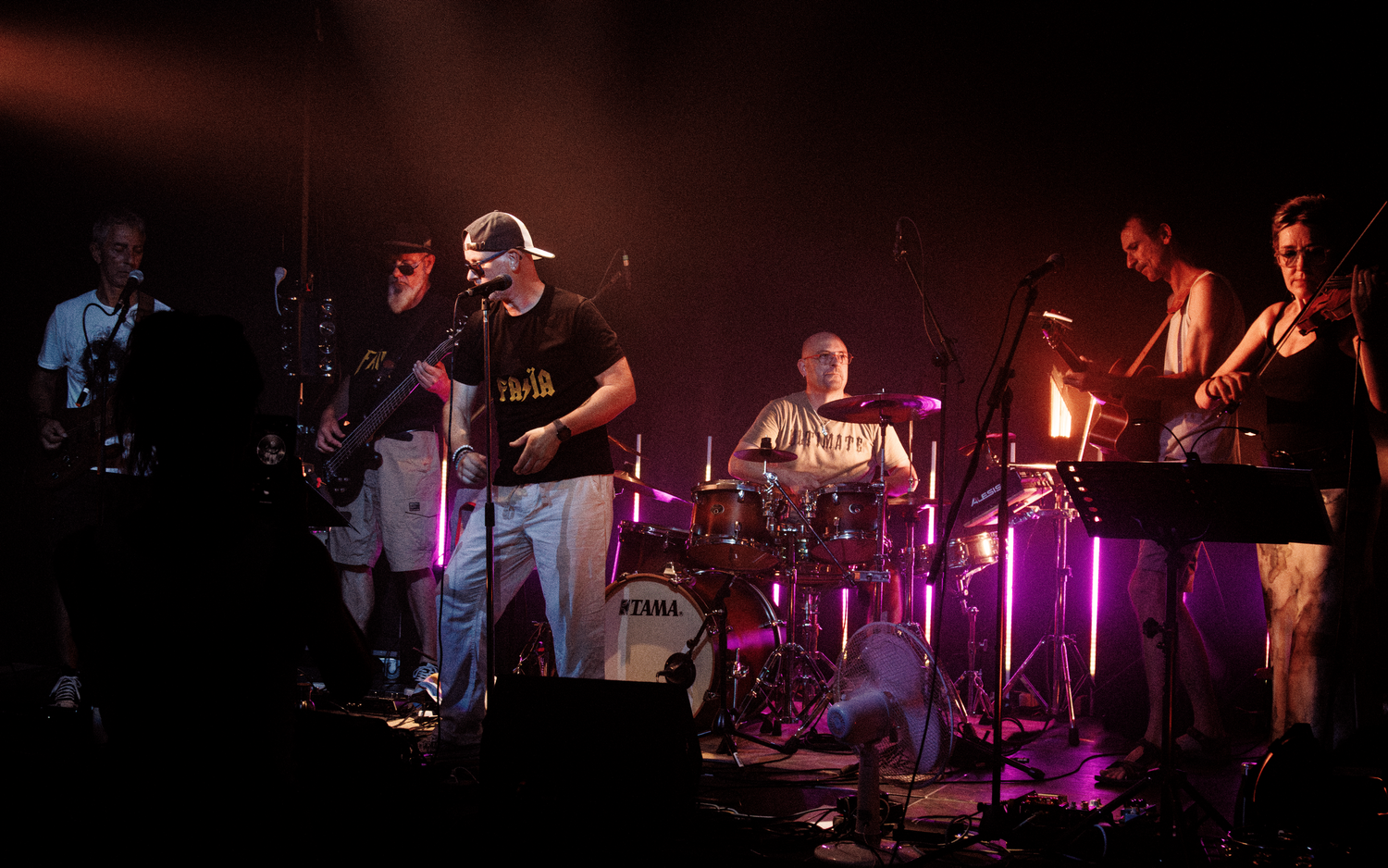 A band performs on stage with colorful lighting and instruments including drums and guitars.