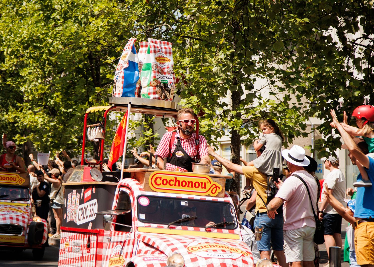 Colorful parade float with Cochonou branding moves through tree-lined street during summer festival.