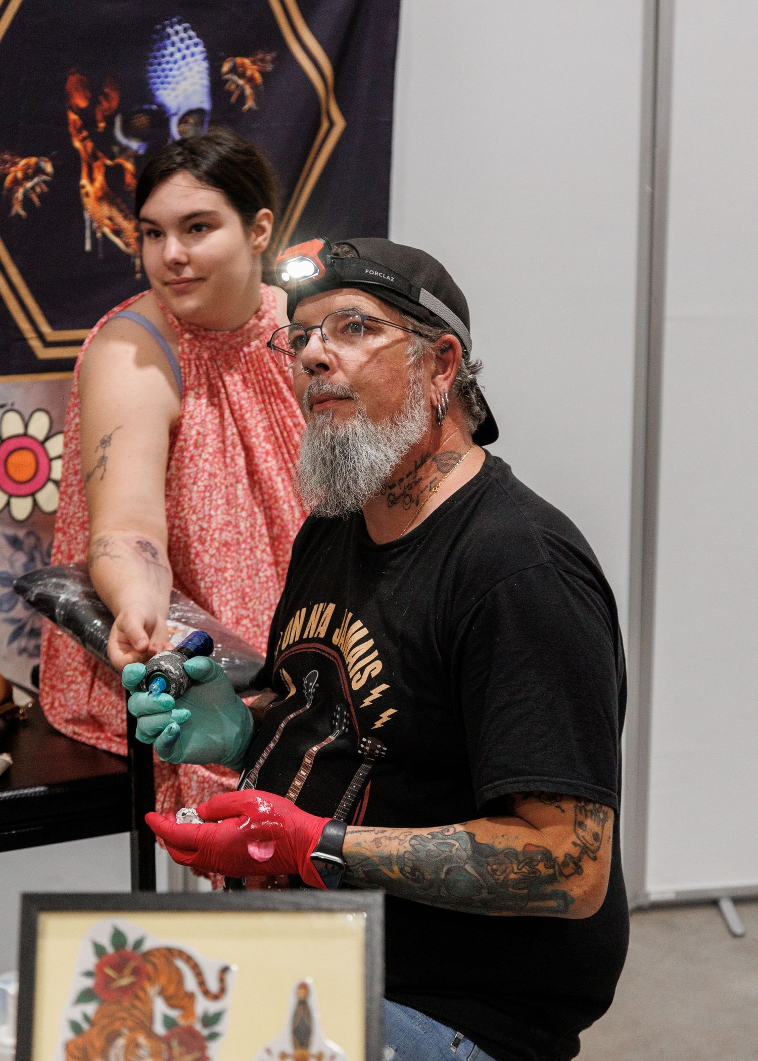 Tattooed artist wearing colorful gloves demonstrates tattooing technique at a convention while a young woman observes nearby.