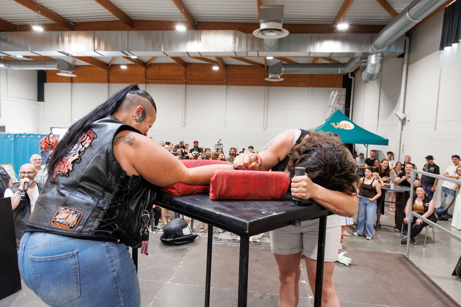Two people arm wrestling on a red padded table at an indoor event, surrounded by a crowd of spectators.