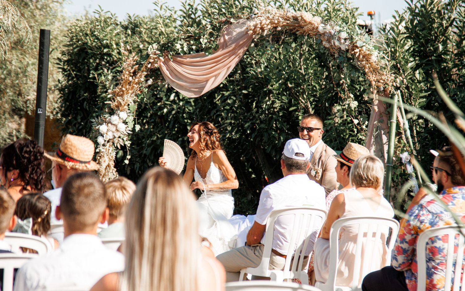 Wedding guests dressed in white gather outdoors for a celebration under a rustic canopy with pink drapery.